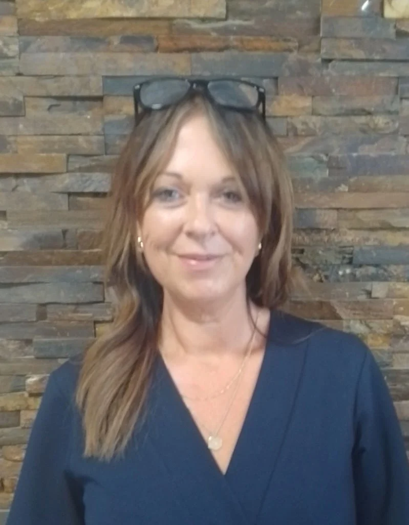 A woman with shoulder-length brown hair, wearing glasses on her head, a navy blue top, and gold necklaces, stands smiling in front of a stone wall.
