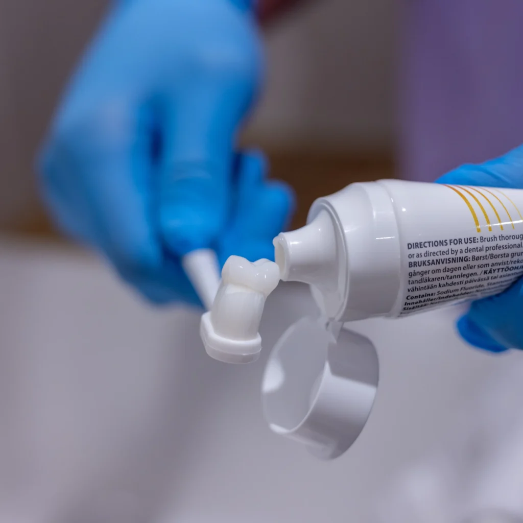A person wearing blue gloves squeezes white toothpaste from a tube onto a toothbrush.