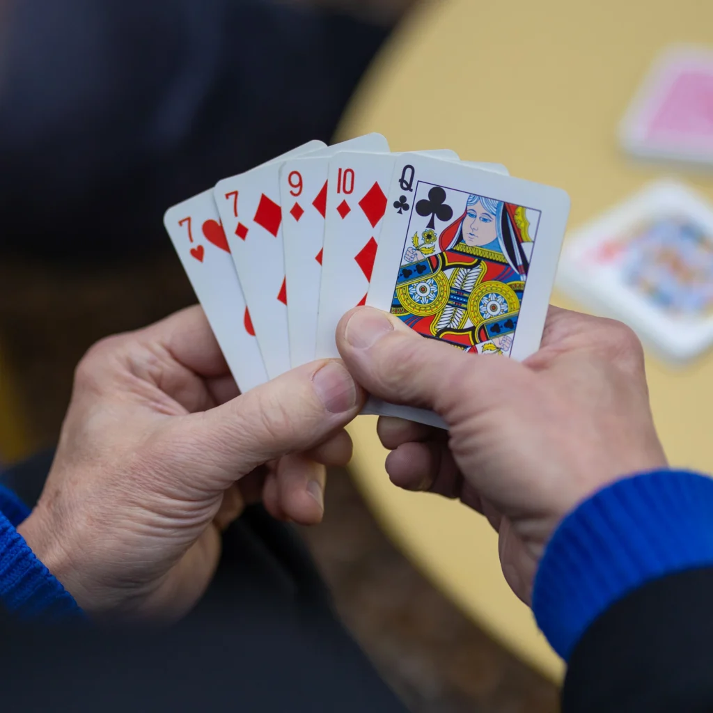 A person holding playing cards, showing the 7, 8, 9, and 10 of diamonds and the queen of clubs, with a blurred table and more cards in the background.