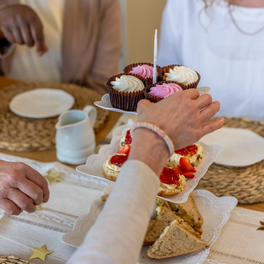 A close-up of a hand reaching for cupcakes on a tiered tray with scones and sandwiches. Three people are seated at a table set for tea with woven placemats and white crockery.