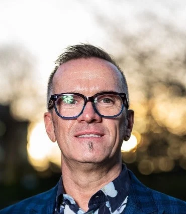A man with short hair, glasses, and a small goatee smiles slightly whilst standing outdoors. He is wearing a patterned shirt and blazer, with blurred trees and sunlight in the background.