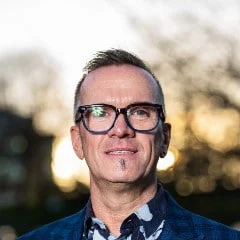 A man with short hair, glasses, and a small goatee smiles outdoors. He is wearing a patterned shirt and a blue jacket, with a blurred background of trees and sunlight.