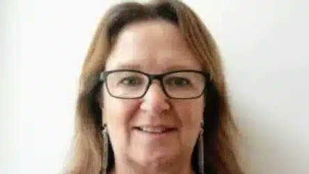 A woman with long brown hair, wearing glasses and earrings, is smiling in front of a plain white background.