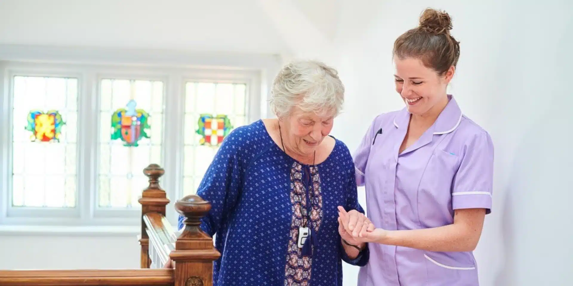 A smiling nurse in a lavender uniform assists an elderly woman in a blue dress as they walk together indoors near a wooden bannister and stained glass windows.