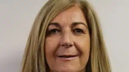 A middle-aged woman with straight, shoulder-length blonde hair is facing the camera and smiling slightly against a plain light background.