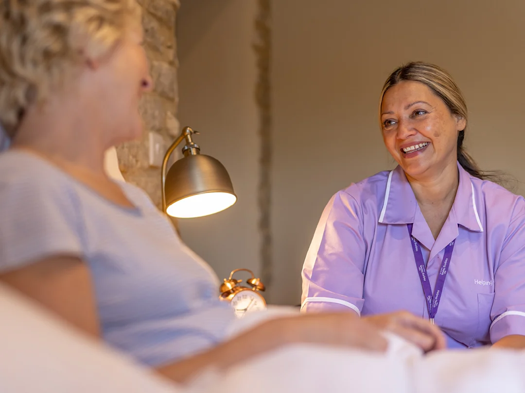 A nurse in a purple uniform sits beside a smiling woman in bed, engaging in a friendly conversation in a cosy room with a lamp and an alarm clock on the bedside table.
