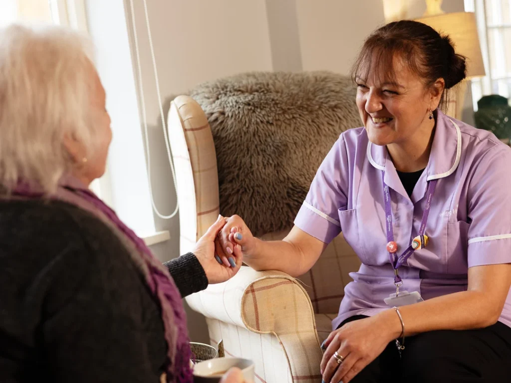 A carer in a purple uniform smiles and holds hands with an elderly woman, providing comfort and support in a cosy, well-lit living room.