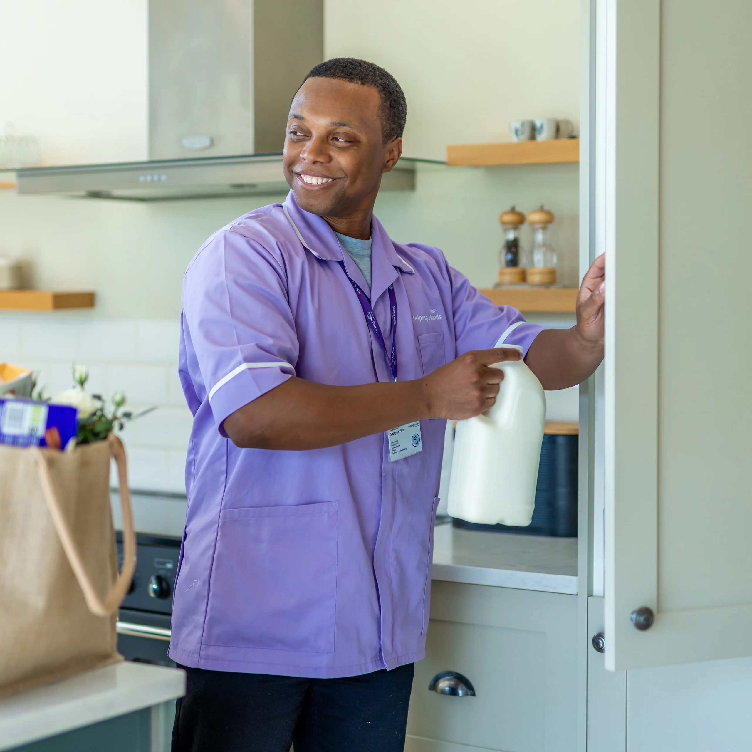 A smiling man in a light purple uniform holds a milk bottle while opening a kitchen cupboard. A shopping bag sits on the counter behind him.