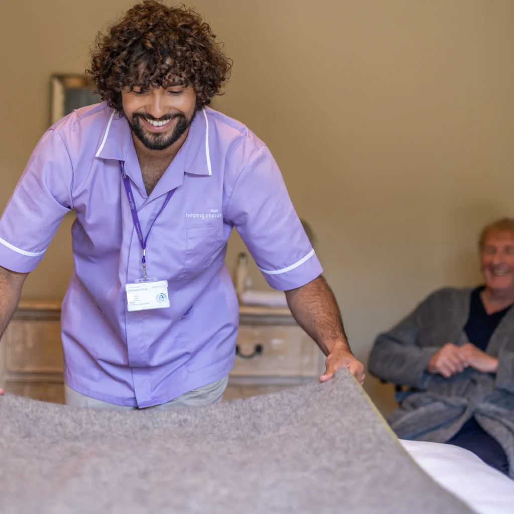 A smiling carer in a purple uniform is making a bed, while an older man in a dressing gown sits on a chair in the background, watching and smiling.