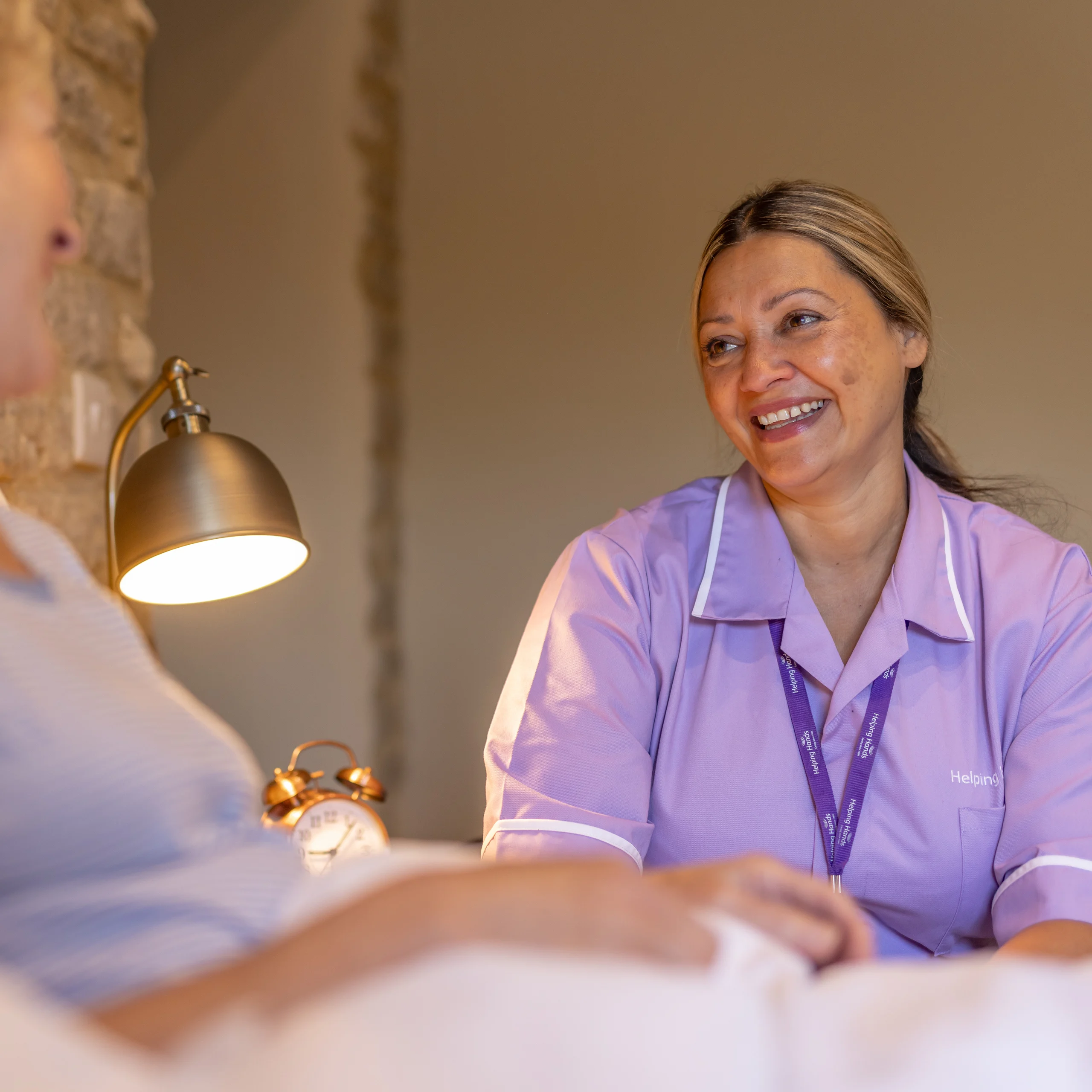A carer in a purple uniform smiles warmly at a person in bed, creating a comforting and supportive atmosphere in a softly lit room.