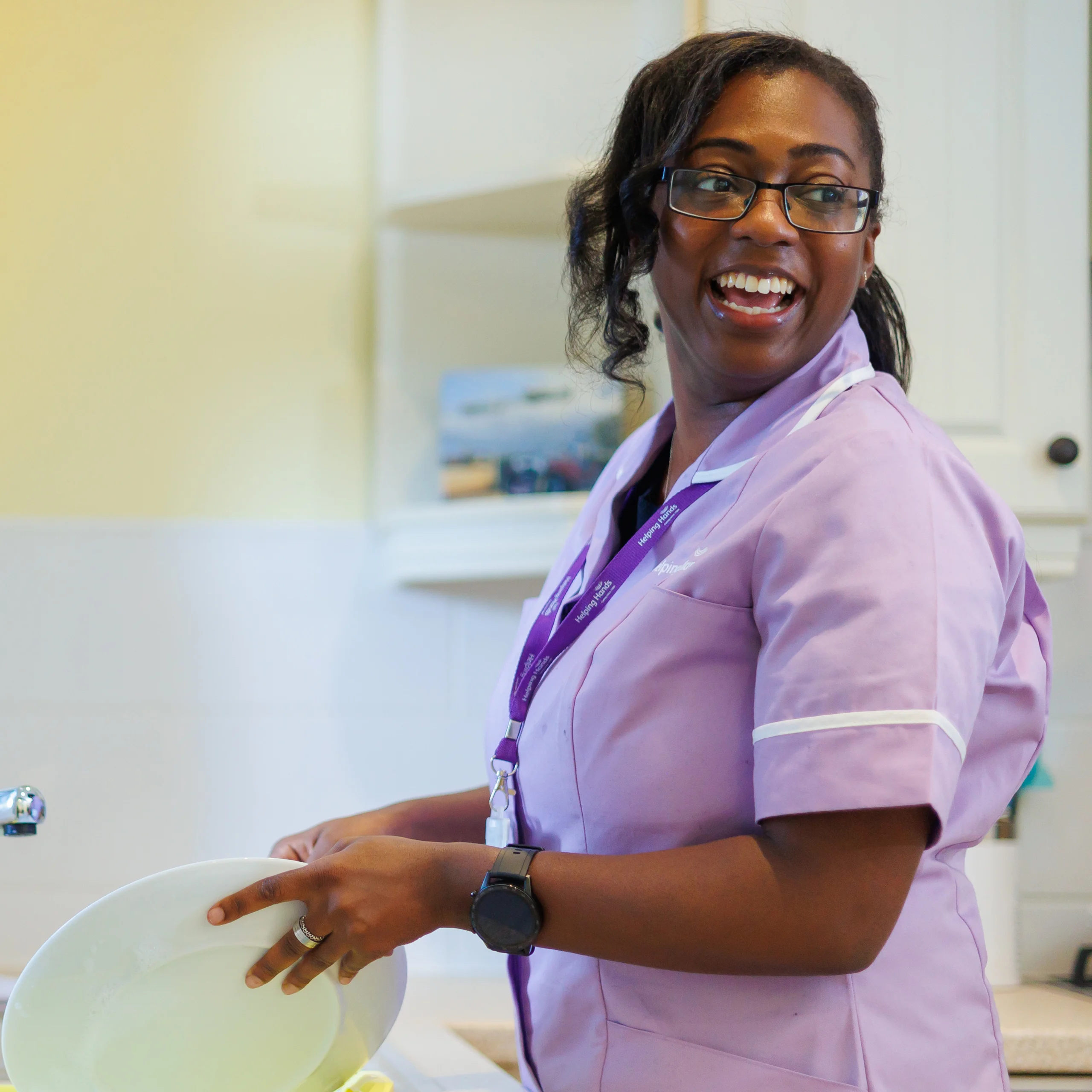A woman wearing glasses and a light purple uniform smiles whilst washing dishes in a bright kitchen.