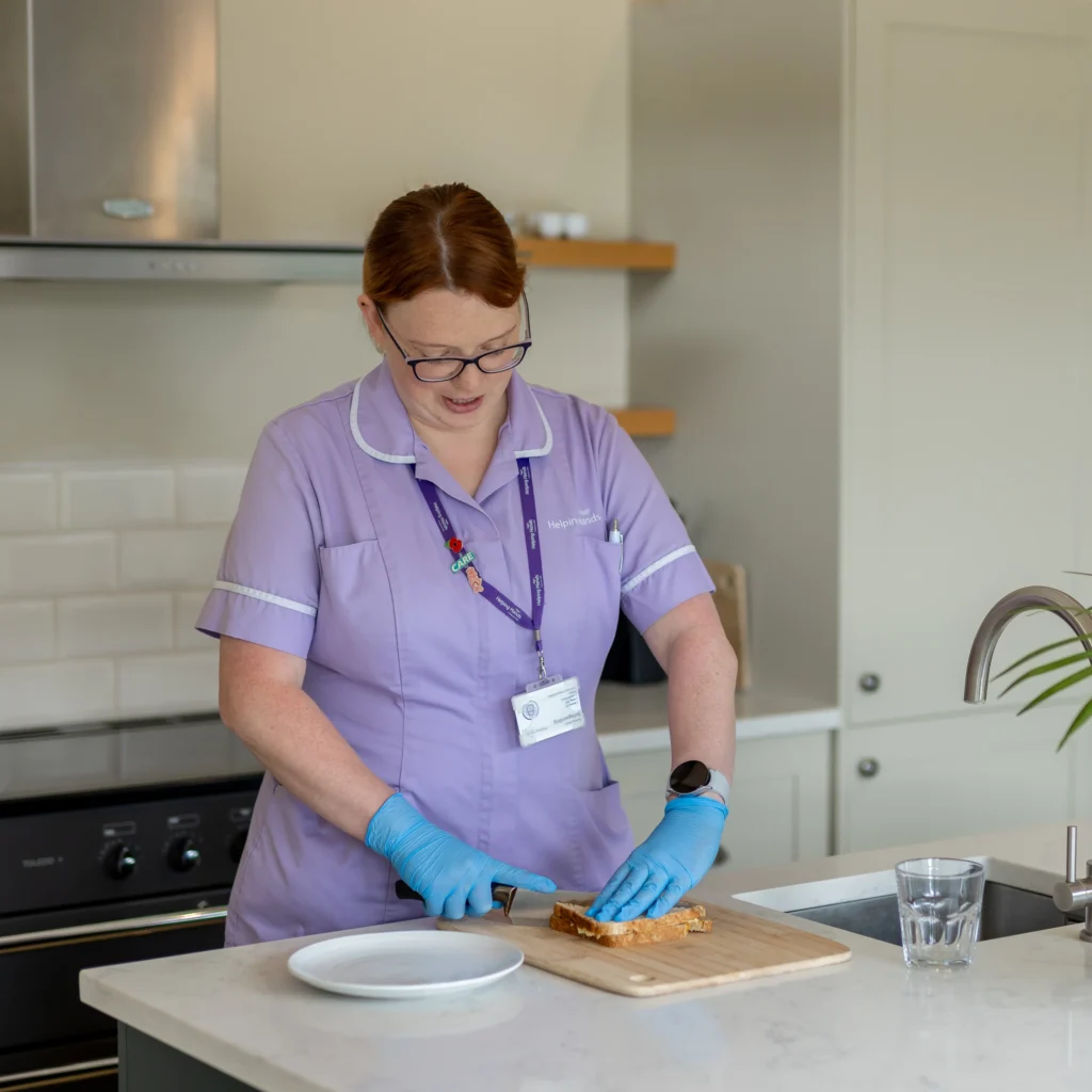 A woman in a light purple uniform and blue gloves slices bread on a wooden chopping board in a modern kitchen. A white plate and a glass of water are on the worktop.