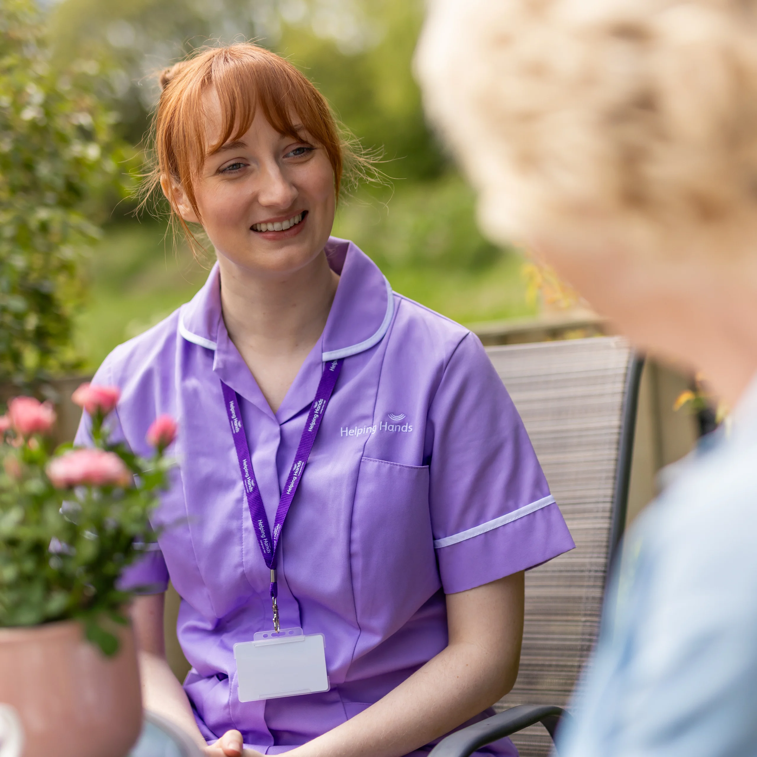 A smiling carer in a purple uniform sits outdoors, speaking with an elderly person. There is a potted plant with pink flowers on the table between them.