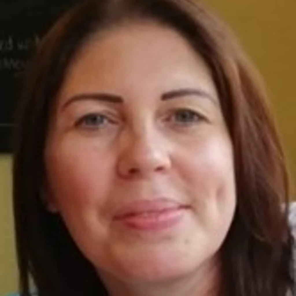 A woman with straight brown hair and light skin smiles gently at the camera against an indoor background.