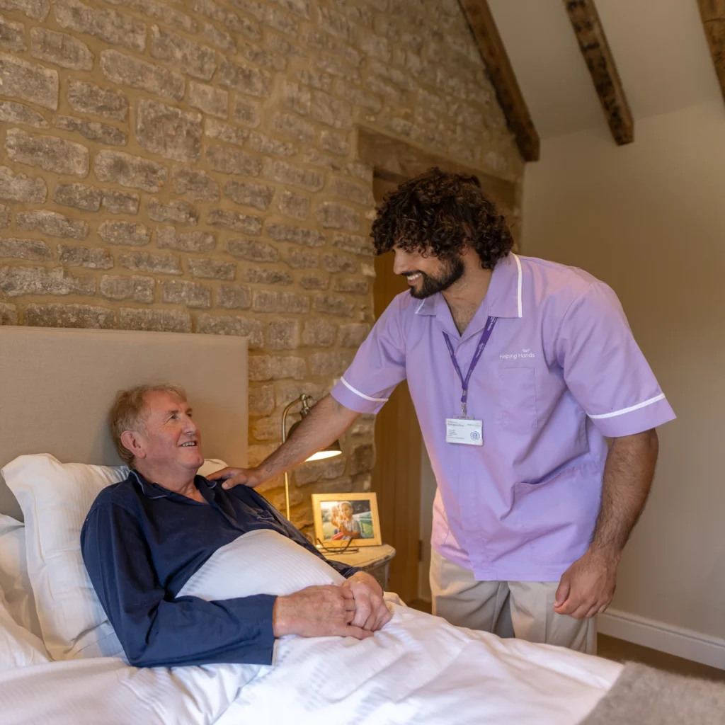 A carer in a purple uniform smiles and chats with an elderly man lying in bed in a cosy, rustic room. The carer rests a hand on the man's shoulder, and a framed photo sits on the bedside table.
