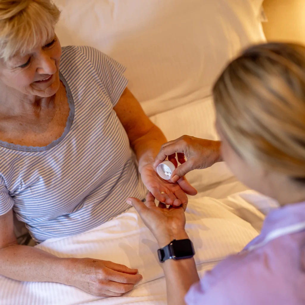 A nurse places white tablets into the palm of an elderly woman who is sitting up in bed, smiling and wearing a striped top, in a warmly lit room.