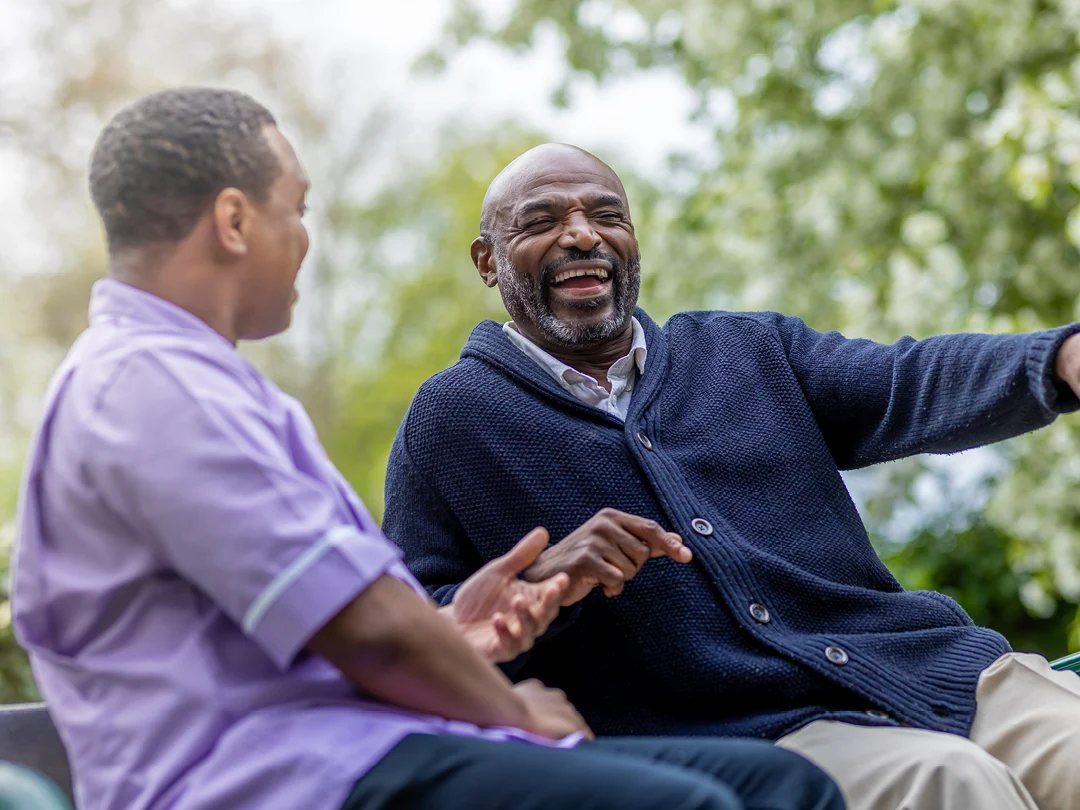 Two men sit outdoors, smiling and laughing together. One man wears a navy cardigan and gestures animatedly, while the other wears a light purple shirt. Trees and greenery are visible in the blurred background.