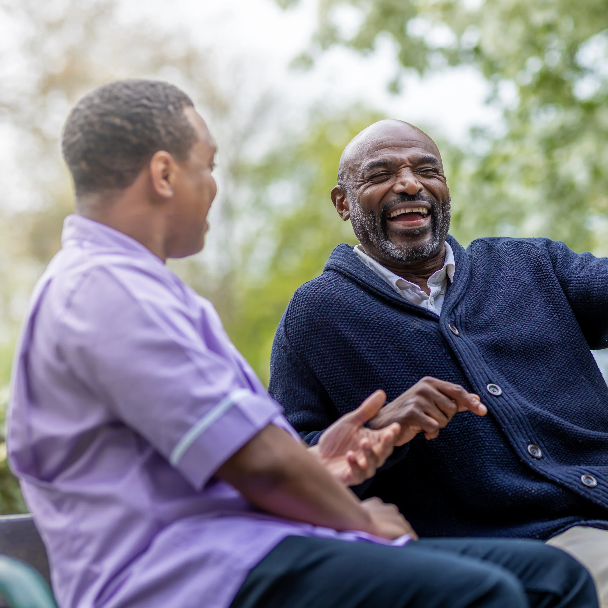 Two men sit outdoors, smiling and laughing together. One wears a dark jumper, the other a light purple shirt. Trees and greenery are visible in the blurred background, suggesting a park or garden setting.