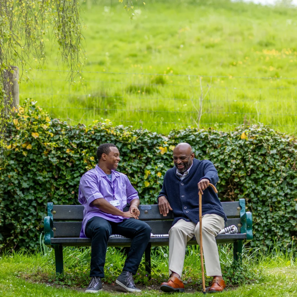 Two men sit on a park bench, smiling and talking. One wears a purple shirt and lanyard, the other has a walking stick, dark jumper, and brown shoes. Green grass and ivy create a lush background.