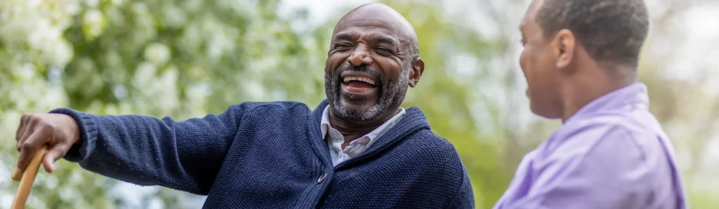 Two men outdoors smiling and laughing together; one man, wearing a dark cardigan and holding a walking stick, is facing the camera, while the other, in a light purple shirt, is seen from the side. Trees and greenery are in the background.