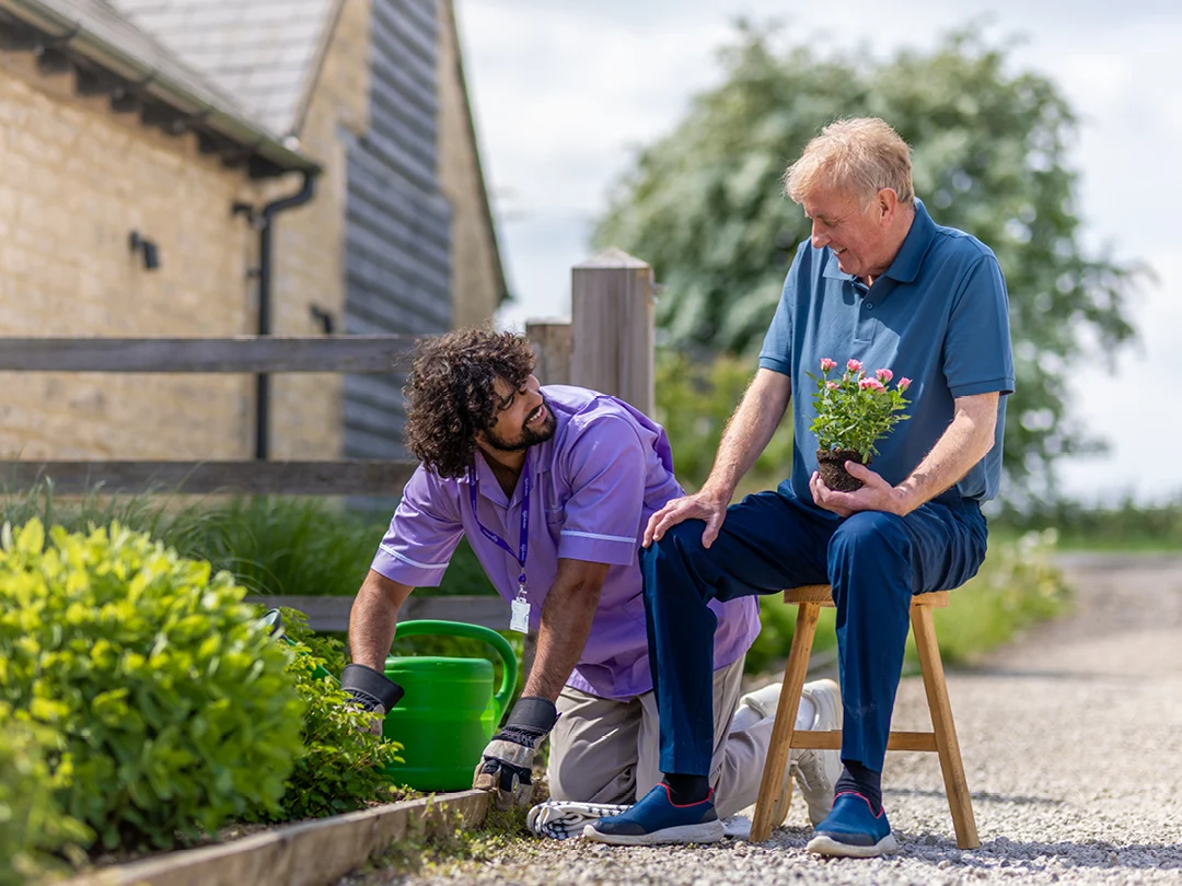 A carer kneels while watering garden plants with a green watering can, as an older man sits on a stool nearby holding a small potted flower. Both are outdoors beside a house on a sunny day.