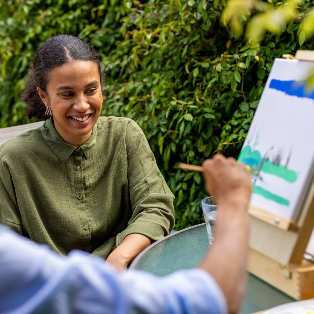 A woman with curly hair and a green shirt smiles while watching someone paint on a canvas at an outdoor table surrounded by greenery.