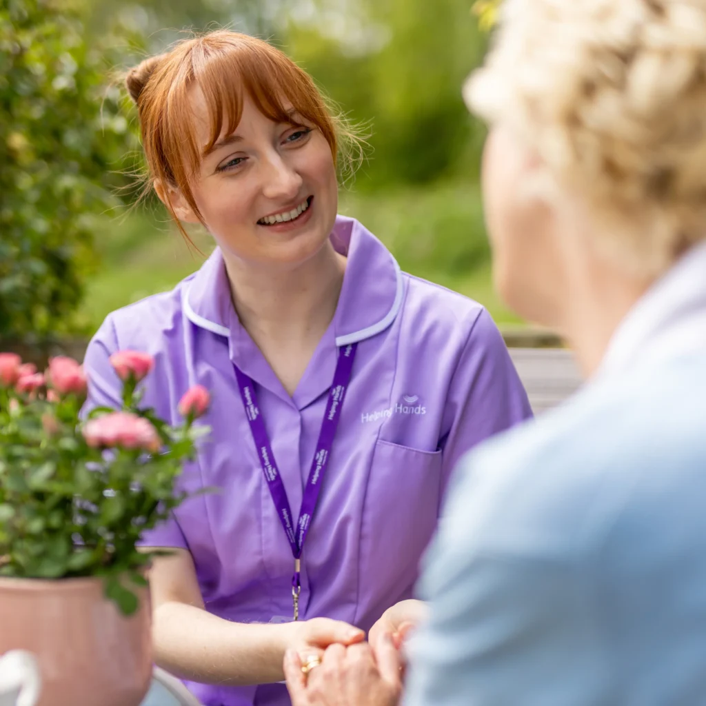 A smiling carer in a purple uniform is holding hands with an older person outdoors, next to a pink potted plant, with greenery in the background.