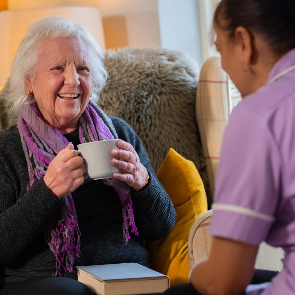 An elderly woman smiles whilst holding a mug, sitting on a sofa with a book on her lap. She faces a person in a purple uniform, suggesting a friendly conversation in a cosy home setting.