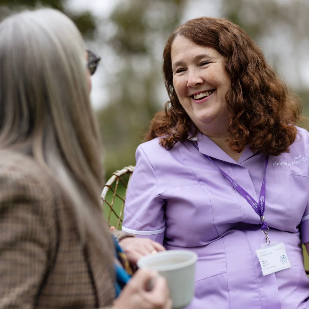 A smiling woman in a lavender uniform with a name badge and lanyard sits outdoors, talking to another woman with long grey hair who is holding a cup. Trees are blurred in the background.