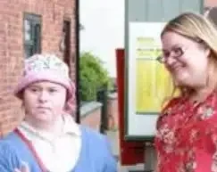 Two women stand outdoors. One wears glasses and a red floral top, smiling, while the other wears a pink hat and blue jumper, looking to the side. They appear to be near a brick building and a yellow sign.