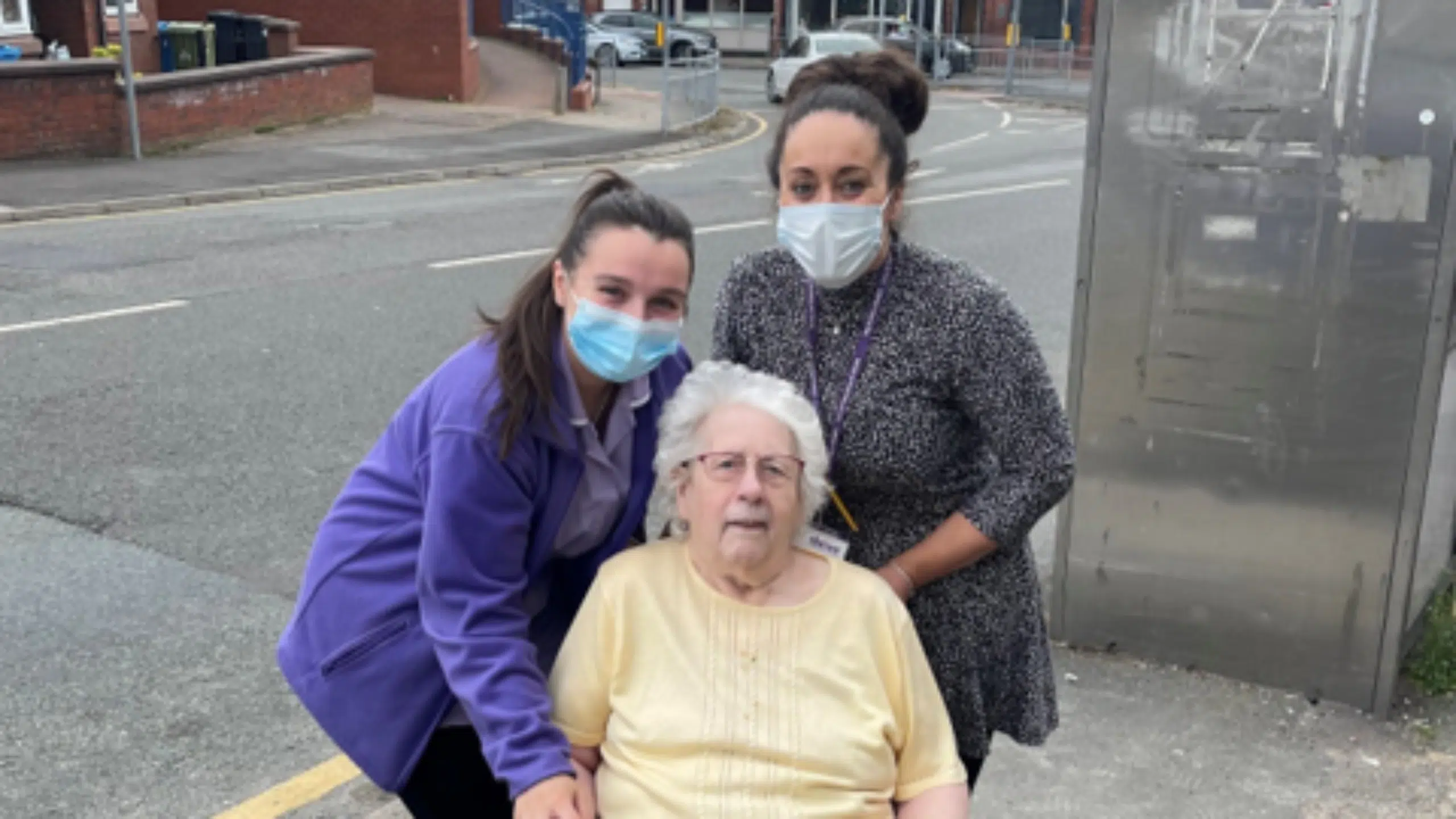 Two women wearing face masks stand next to an elderly woman in a wheelchair; they are outdoors on a pavement near a road, posing together for the photo.