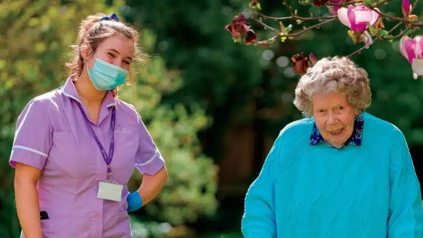 A smiling elderly woman in a blue jumper walks outdoors with a walking stick, accompanied by a female carer in a purple uniform and face mask. Blossoming flowers hang from a tree branch above them.
