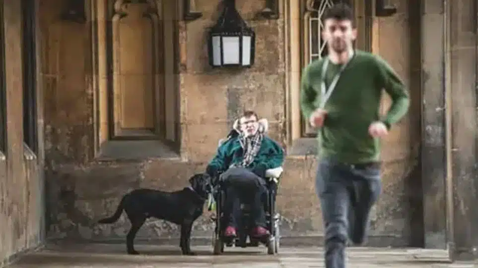 A person in a wheelchair with a black assistance dog sits near an arched building entrance, whilst another person in a green shirt jogs past in the foreground.