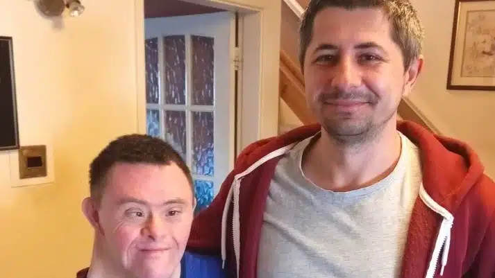 Two men are standing indoors, smiling at the camera. One man has Down’s syndrome and is wearing a blue shirt, while the other man, wearing a red hoodie and grey T-shirt, has his arm around him. They appear to be in a home setting.
