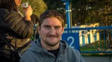 A man with short brown hair and a beard, wearing a grey hoodie, smiles at the camera outdoors with blue fencing and another person holding a drink in the background.