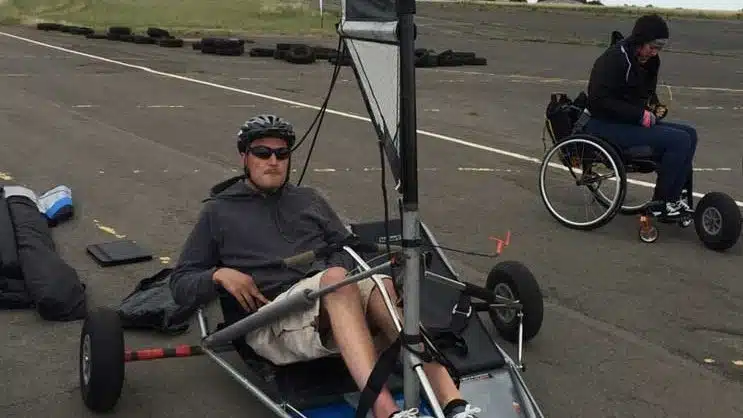 A man wearing a helmet and sunglasses sits in a pedal-powered go-kart on an outdoor track, while another person in a wheelchair is nearby. Tyres and equipment are scattered around on the pavement.