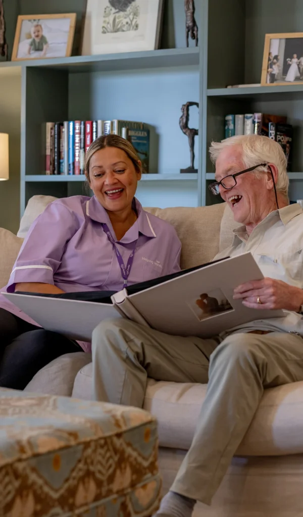 A smiling elderly man and a carer sit on a sofa together, looking through a photo album. They appear happy and engaged, with shelves of books and soft lighting in the background.