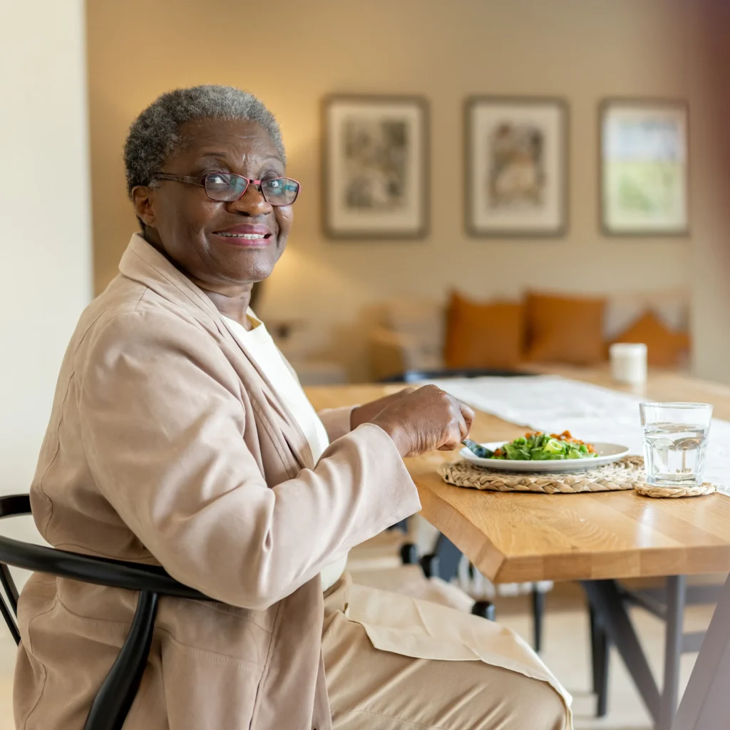 An elderly person with short grey hair and glasses sits at a wooden table, smiling, and eating a salad. A glass of water and framed artwork decorate the modern, cosy room in the background.