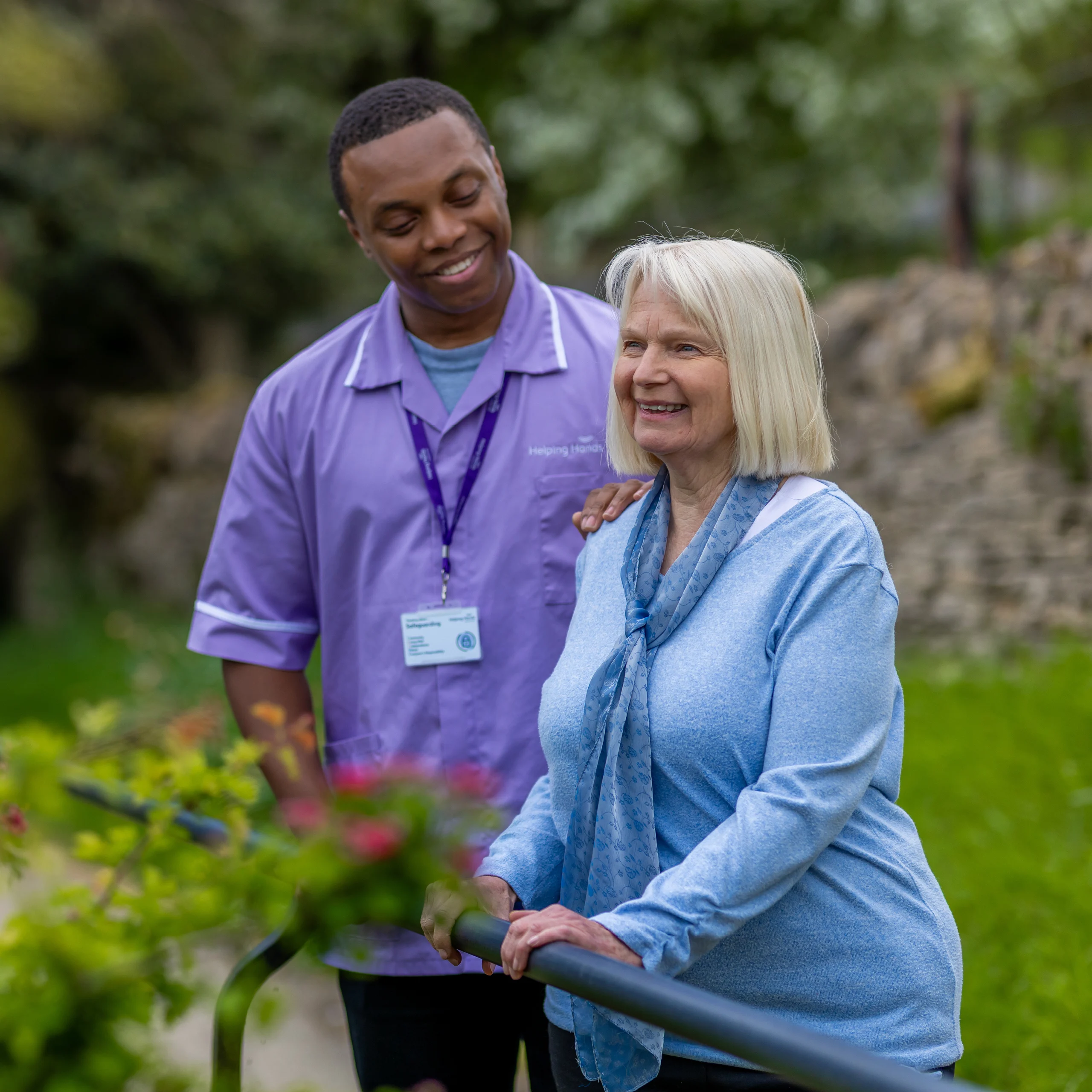 A smiling elderly woman stands outdoors holding a handrail, accompanied by a male carer in a purple uniform who stands beside her, also smiling. The background is green and blurred, indicating a garden or park setting.