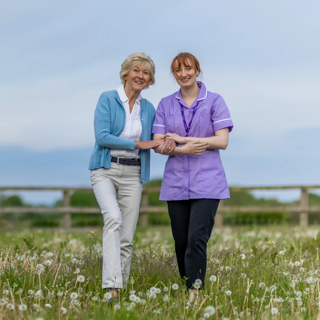 An elderly woman and a carer in a purple uniform walk arm-in-arm through a grassy field with dandelions. Both are smiling, and the background features a wooden fence and a cloudy sky.