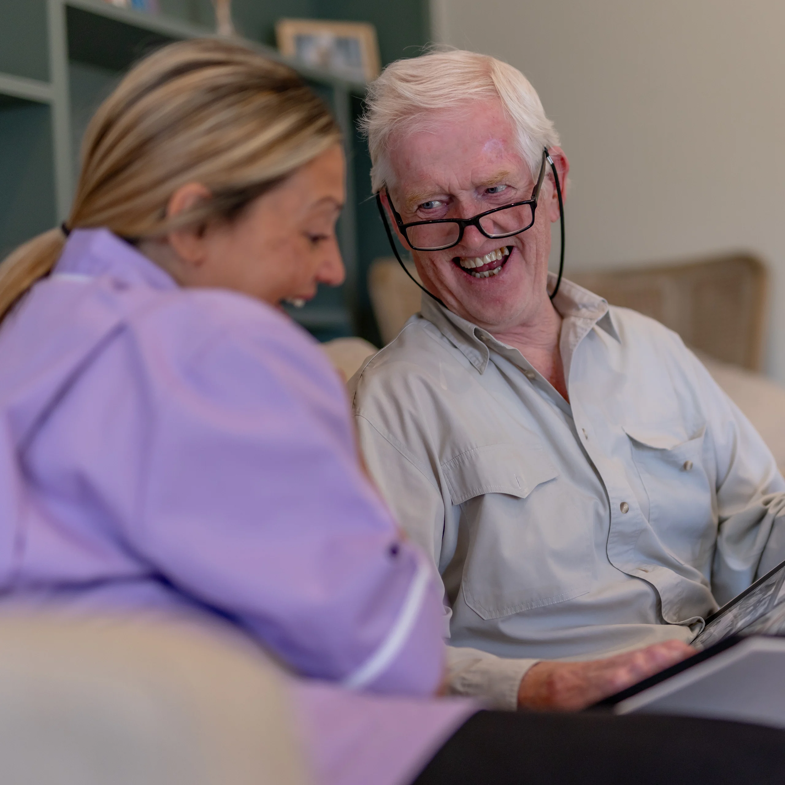 An older man with glasses and a younger woman sit on a sofa, smiling and laughing together while looking at a tablet device. The atmosphere is warm and friendly.