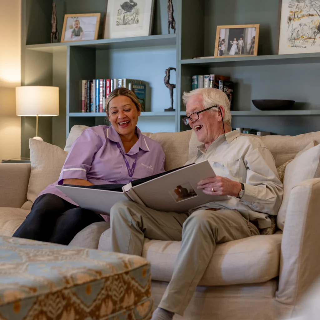 An elderly man and a carer sit on a sofa, smiling and looking at a large photo album together in a cosy, well-lit living room with bookshelves and framed photos in the background.