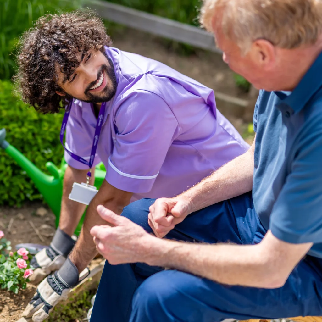 A carer in a purple uniform smiles while gardening with an older man. They sit outdoors near blooming flowers, enjoying a friendly conversation.
