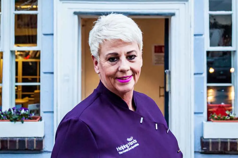 A woman with short white hair, wearing a purple uniform with a Helping Hands logo, stands smiling in front of a building with white-framed windows and a doorway.