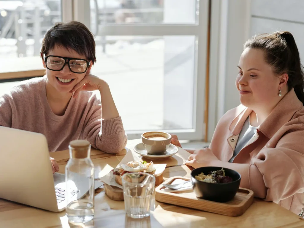 Two women sit at a table by a window, smiling and looking at a laptop. They have food, drinks, and water on the table. Sunlight streams in, creating a warm and cheerful atmosphere.