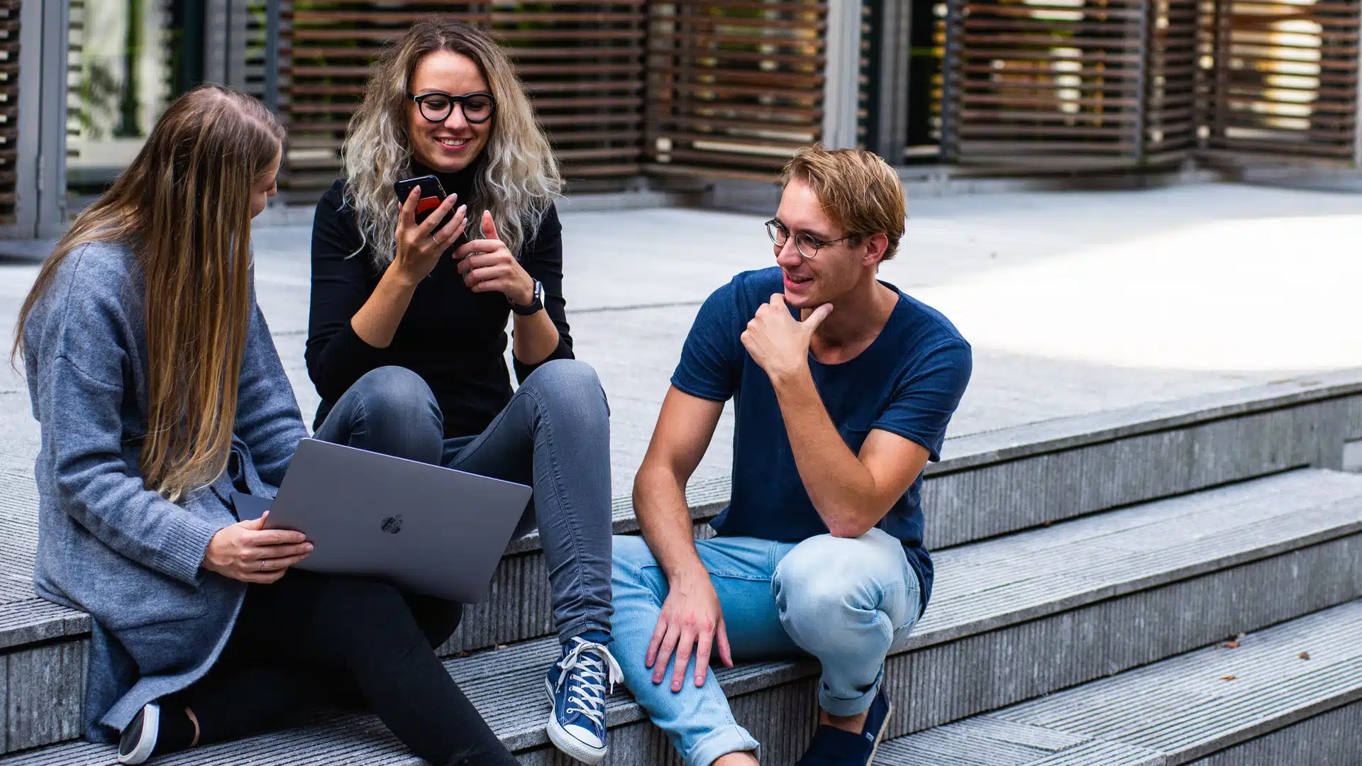 Three young adults sit on outdoor steps. One woman holds a laptop, another laughs while showing something on her mobile, and a man sits nearby smiling. They appear to be enjoying a casual conversation.