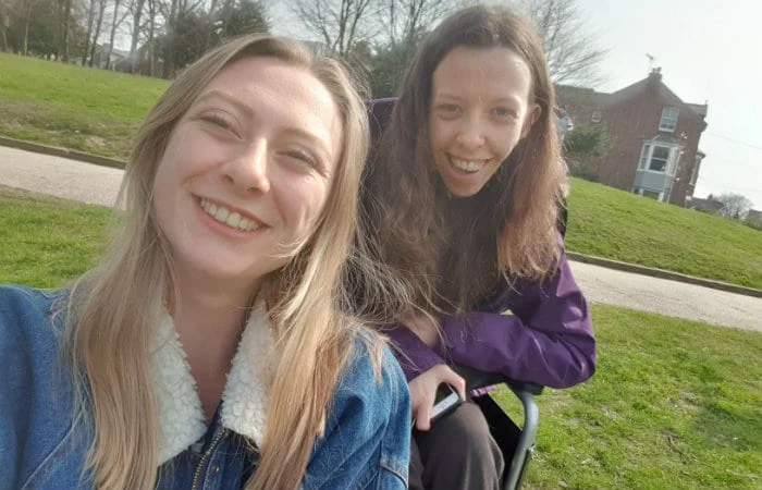 Two young women smiling outdoors in a grassy park, one sitting in a wheelchair and the other standing beside her. Trees and a brick building are visible in the background on a sunny day.