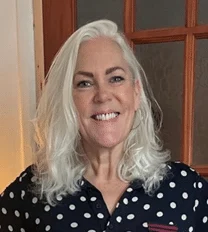 A smiling woman with shoulder-length white hair wears a black shirt with white polka dots, standing indoors in front of a wooden-framed glass door.