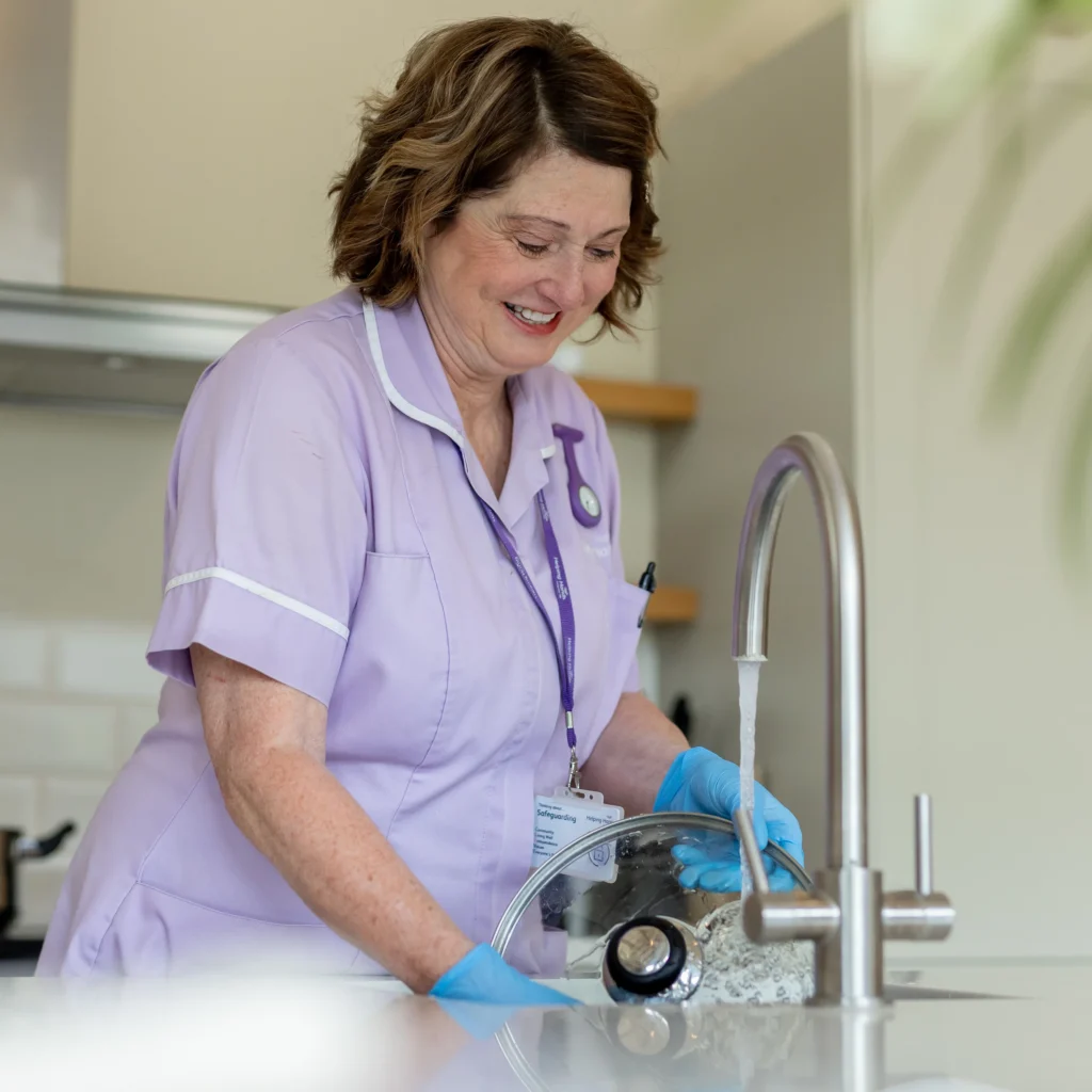 A woman in a light purple uniform and blue gloves smiles while washing a glass lid at a kitchen sink under running water. The background is bright and modern.