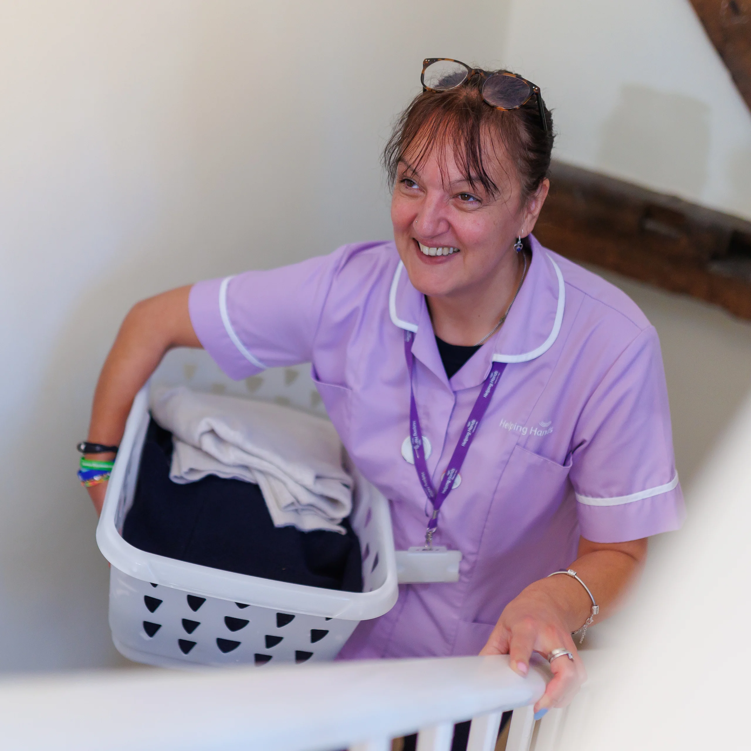 A smiling woman in a purple uniform carries a laundry basket with folded clothes up a staircase. She has glasses resting on her head and wears a lanyard round her neck.